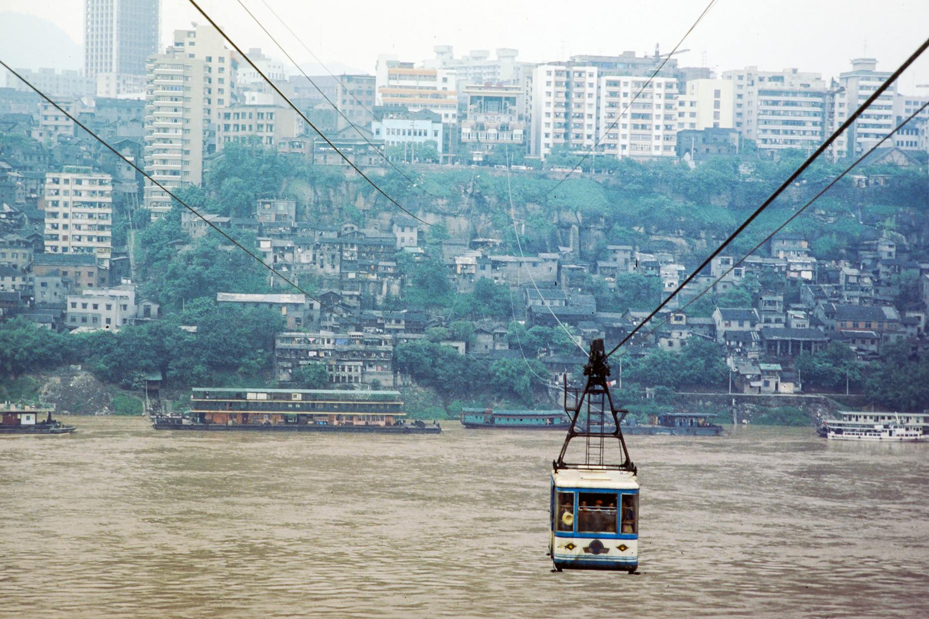 Cable car traversing a river with a cityscape backdrop, blending urban architecture with natural scenery.