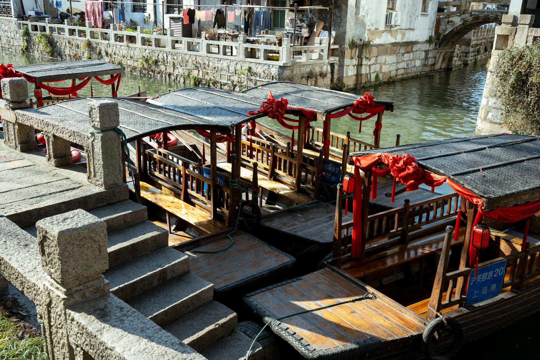 Traditional wooden boats adorned with red fabric moored in a historic Shanghai canal under bright daylight.